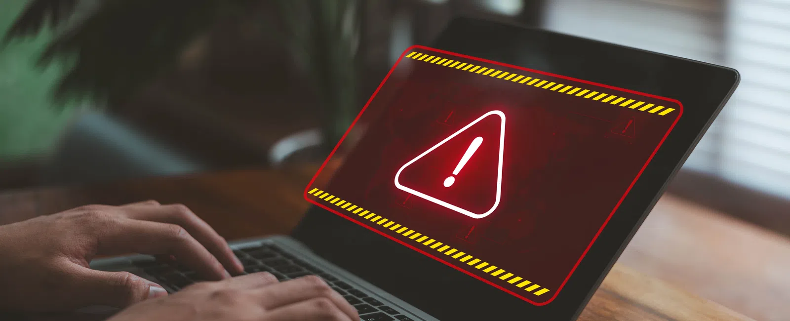 Person typing on laptop showing red warning alert with exclamation mark on screen at home.
