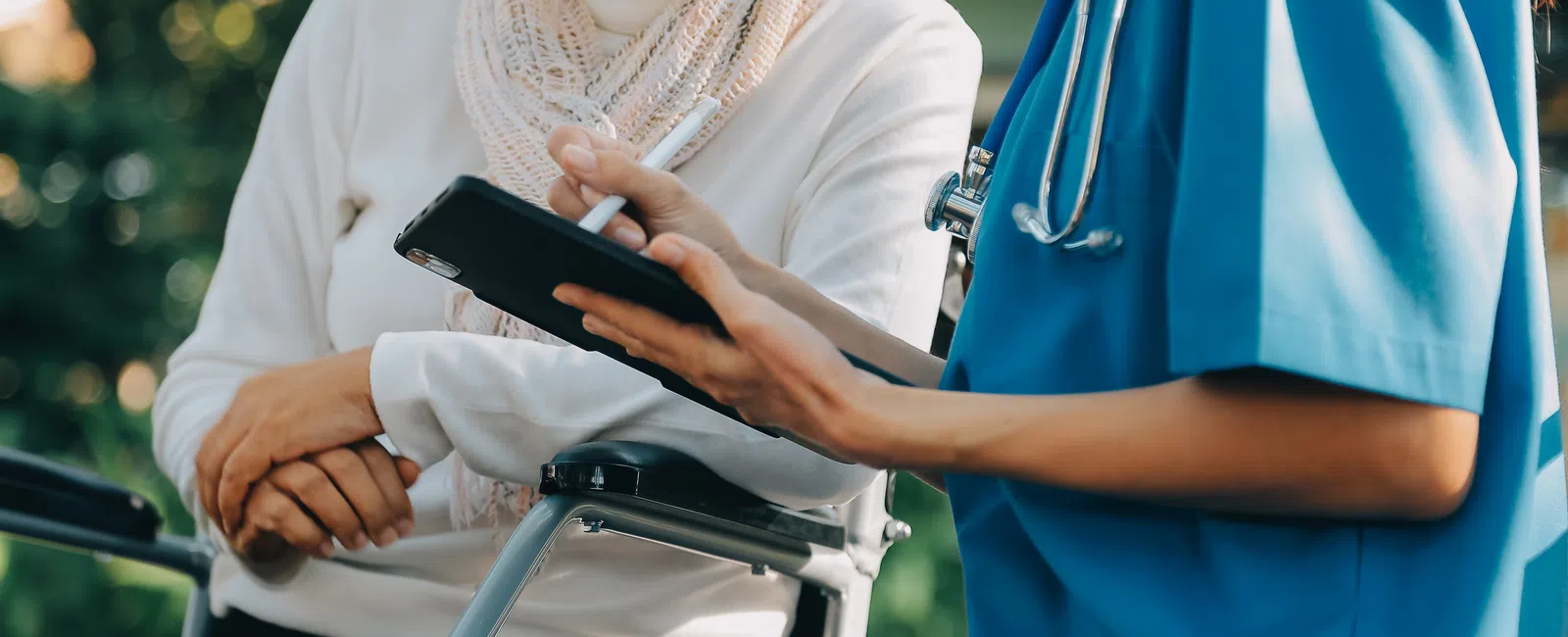 Healthcare professional in blue scrubs consulting an elderly woman in a wheelchair using a tablet outside.
