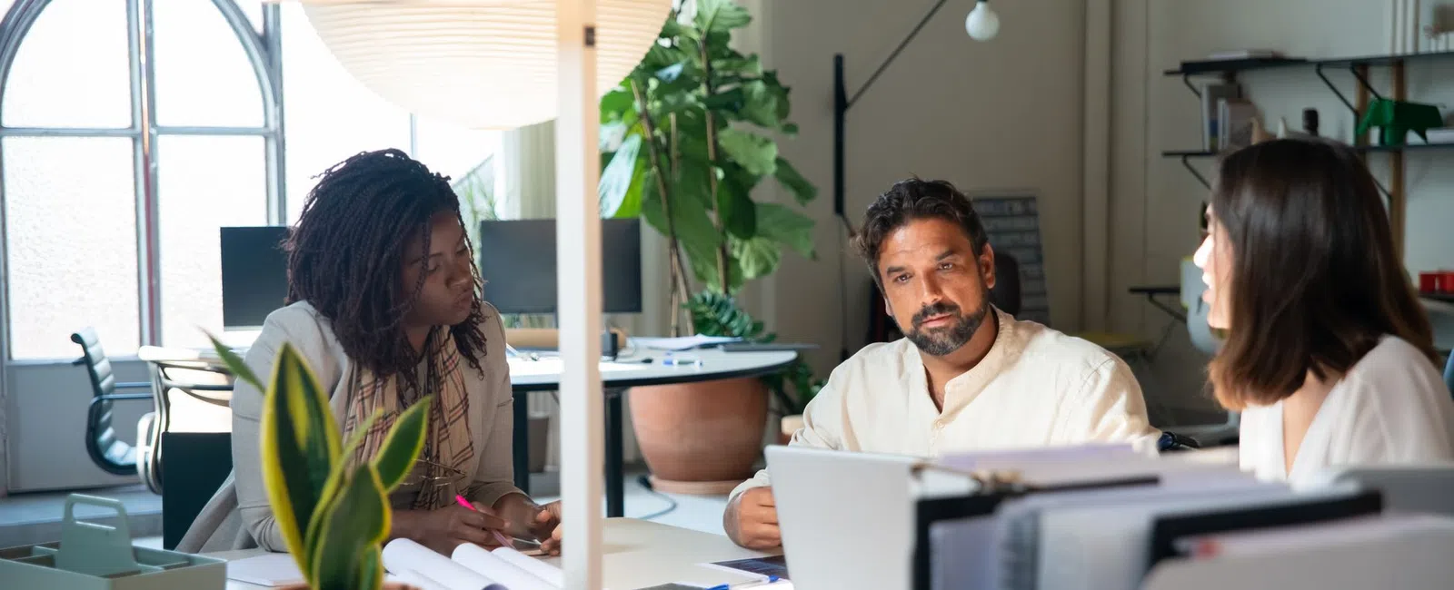 Diverse team in office engaged in a focused discussion around a desk with laptop and documents under a modern lamp.