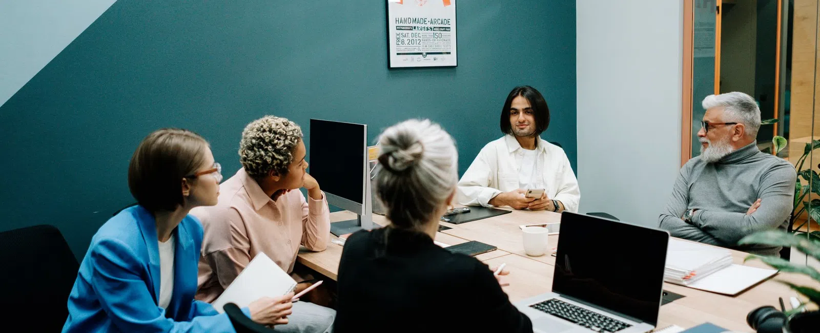 Diverse team having a discussion around a table in a modern office meeting room with laptops and notebooks.