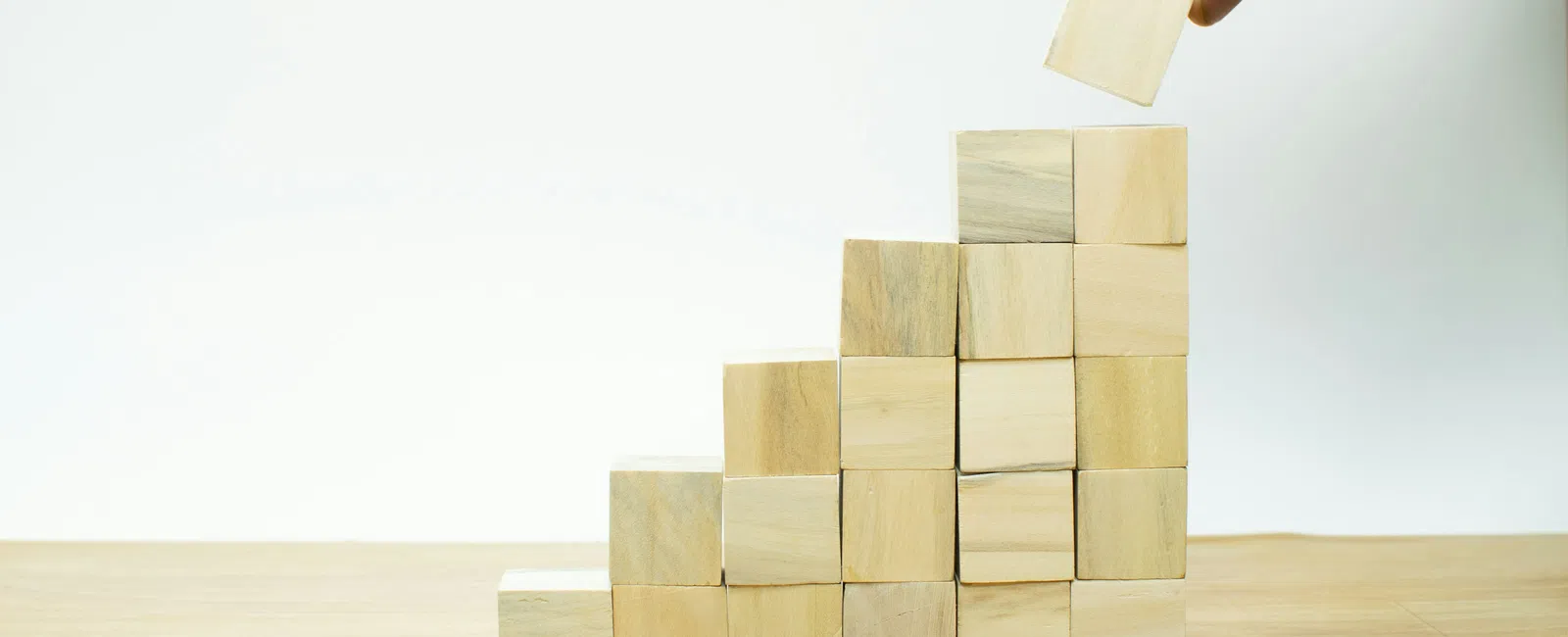 Hand placing a wooden block on top of a staircase-like stack of wooden cubes on a table against white background