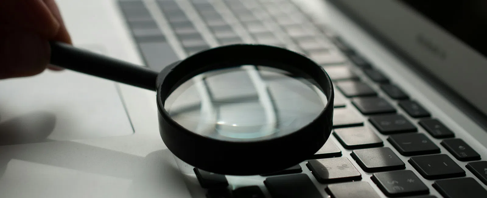 Hand holding magnifying glass over laptop keyboard, highlighting keys and focus on digital search.
