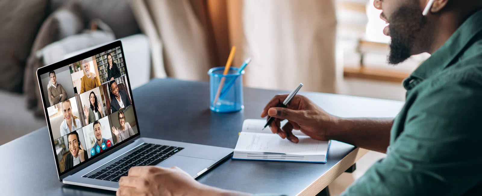 Man wearing glasses and earbuds taking notes during a virtual team meeting on a laptop at home.