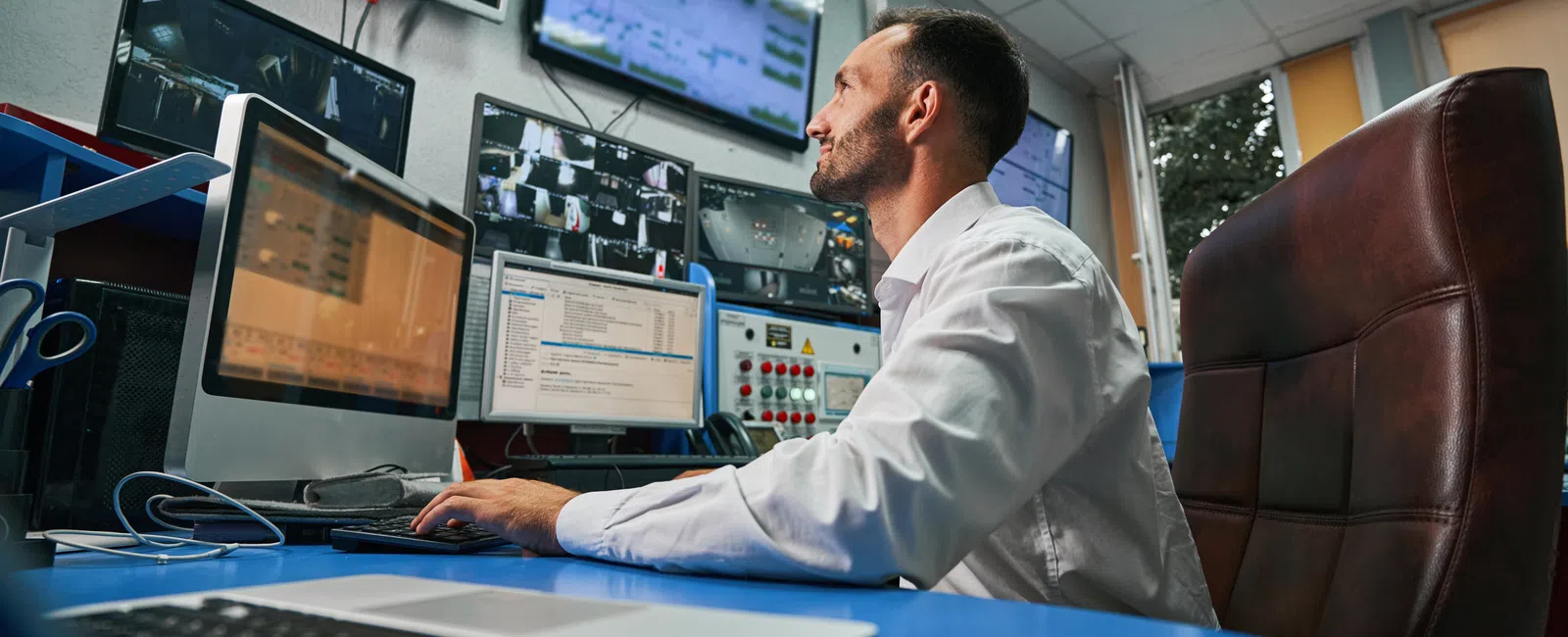 Man monitoring multiple computer screens displaying surveillance footage and data in a control room environment.