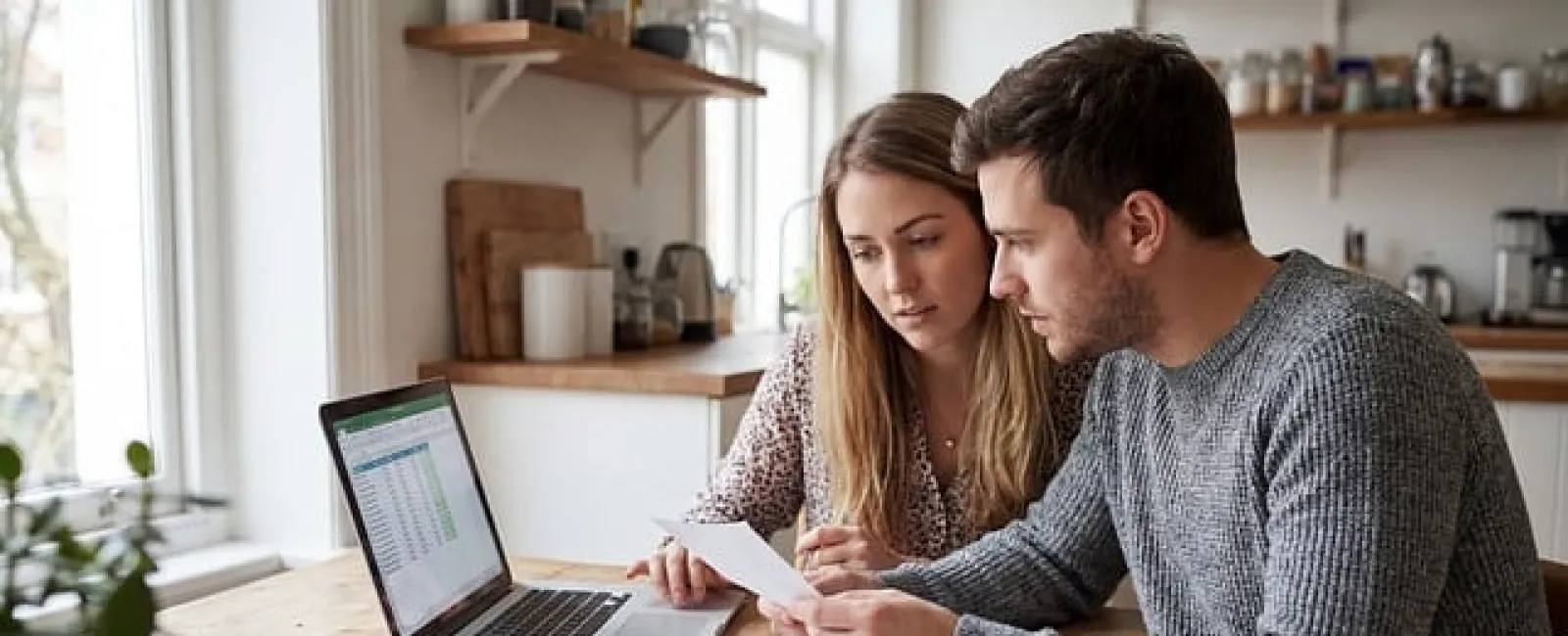 Couple reviewing financial documents and laptop spreadsheet together at wooden kitchen table with coffee cup.