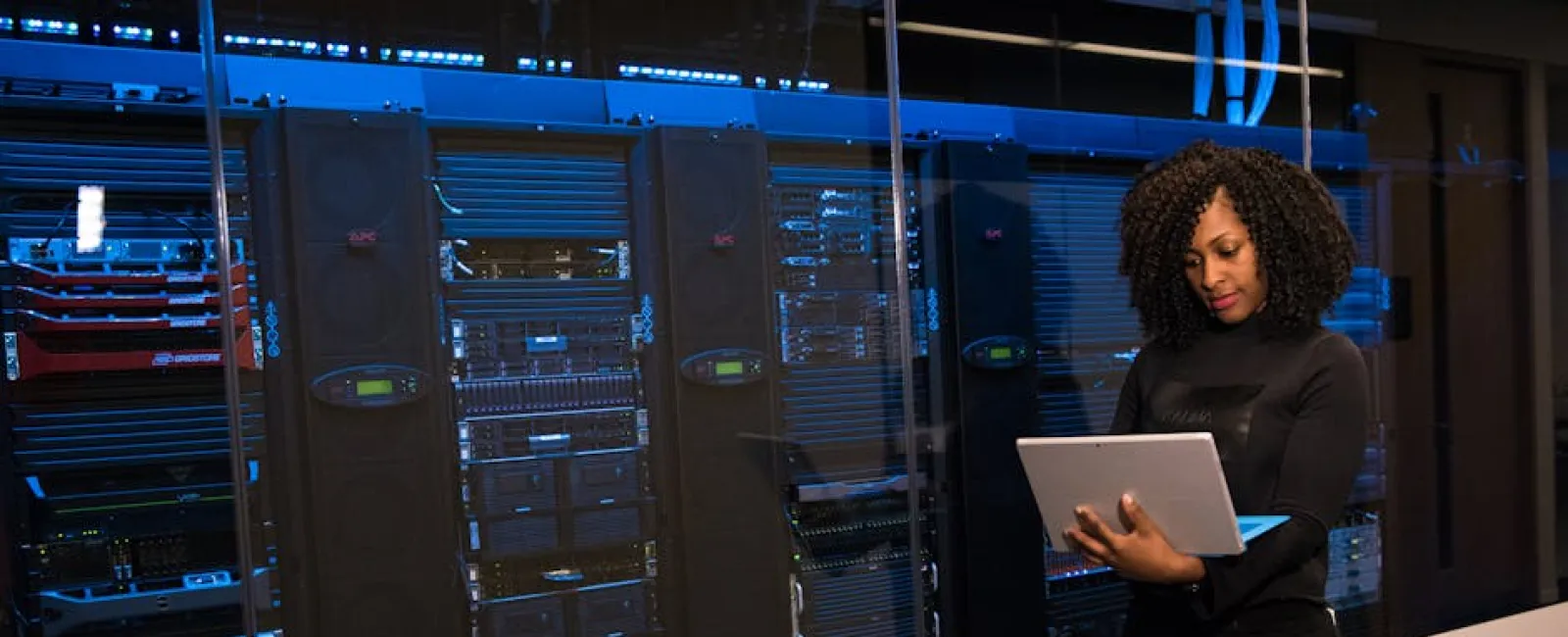 Woman in black outfit holding a laptop in a server room with glass walls and visible server racks.