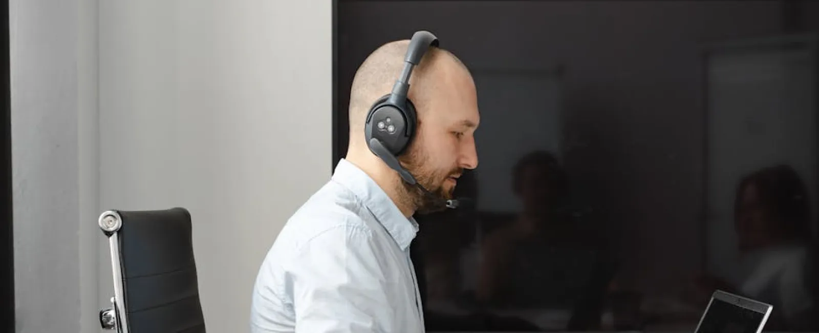 Man with headset working on laptop at desk in modern office environment with documents and smartphone nearby