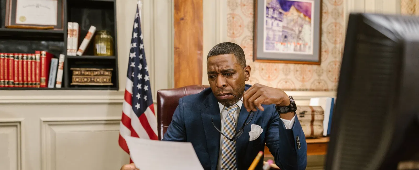 Man in suit reviewing documents at a wooden desk with American flag and computer in government office