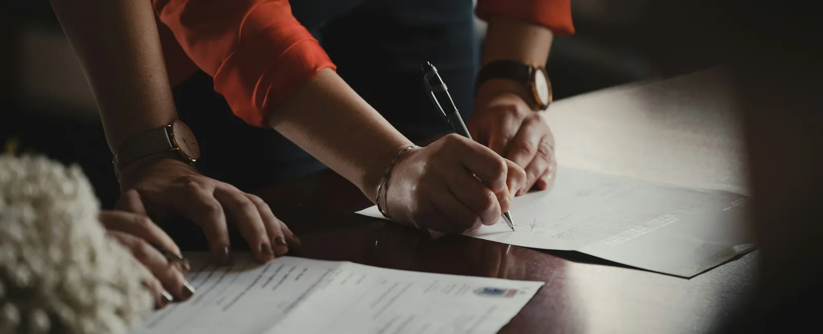 Person in orange shirt signing a document on a wooden table with other papers present