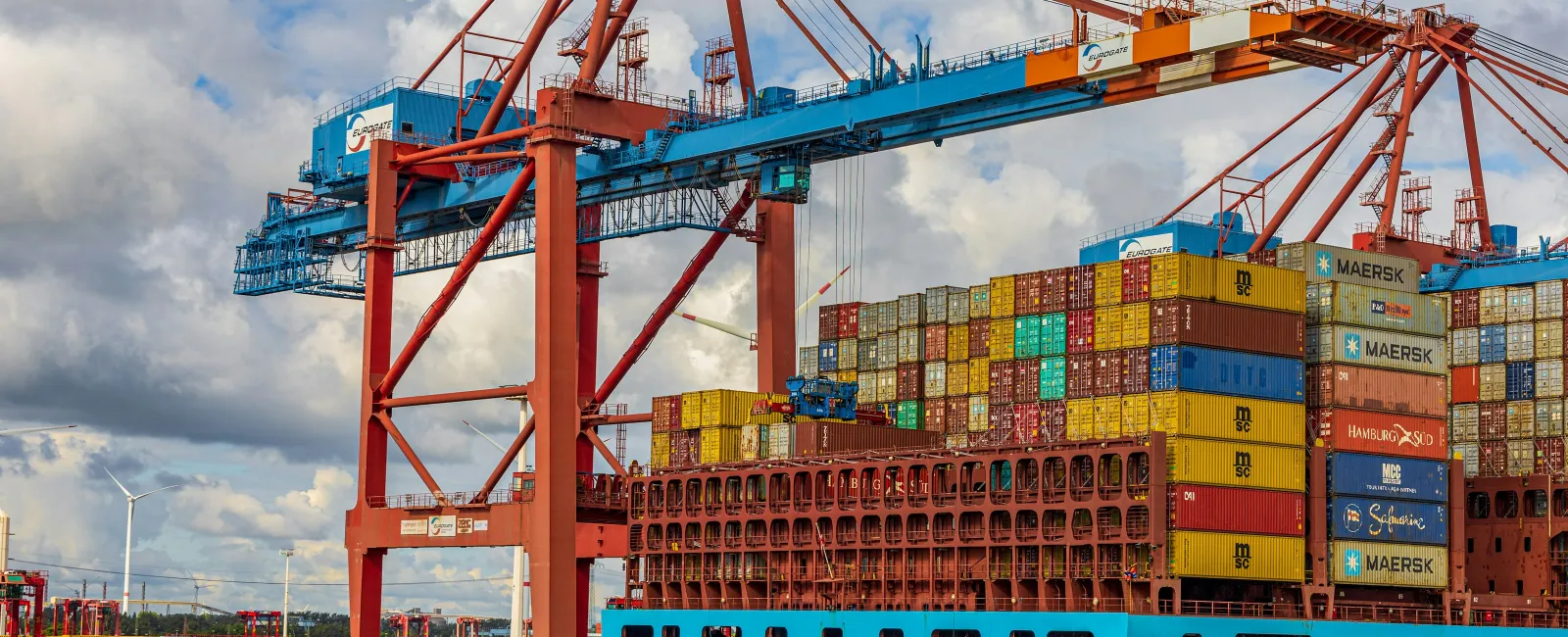 Large cargo ship being loaded with colorful shipping containers by heavy cranes at a busy port under cloudy sky