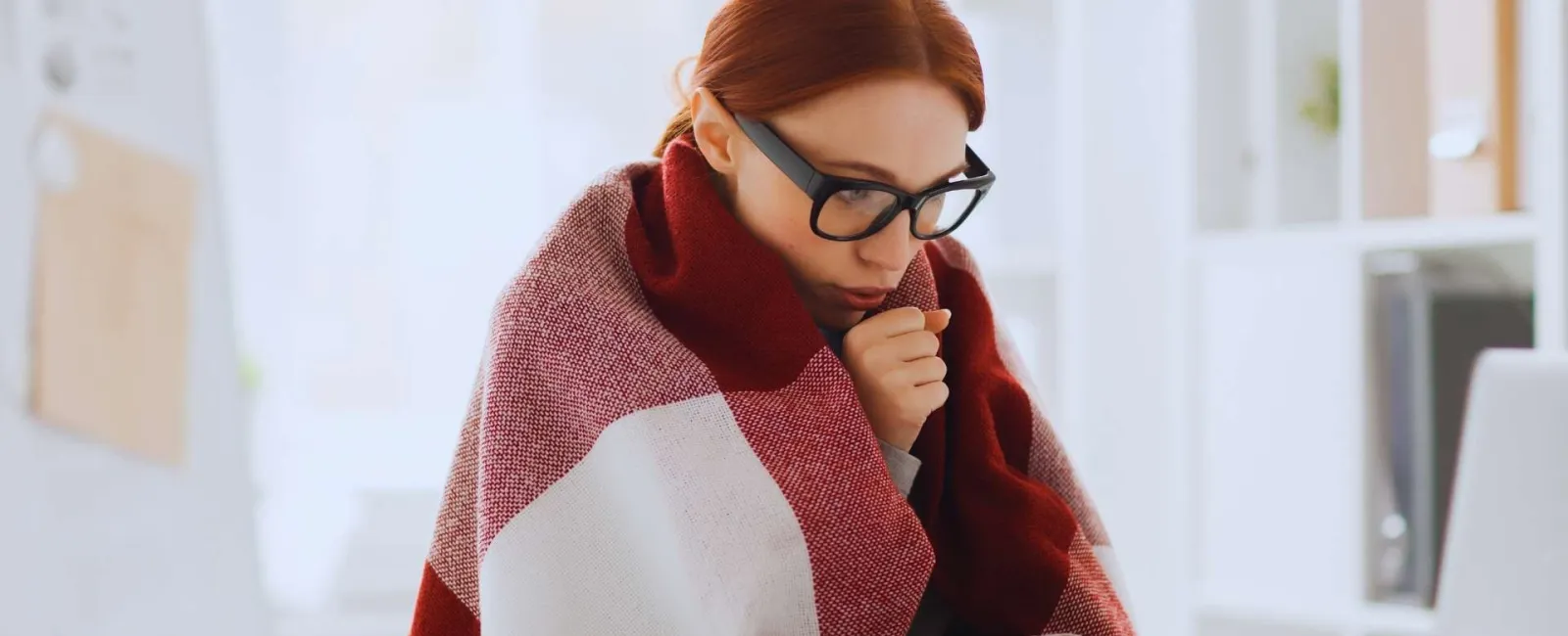 Woman wrapped in a blanket working on a laptop in a bright home office feeling cold.