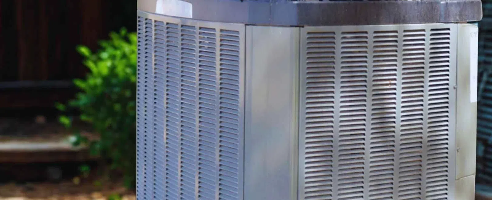 Outdoor central air conditioning unit with metal casing and ventilation grilles in a backyard setting.