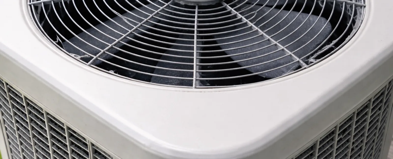 Outdoor home air conditioning unit with metal fan and protective grille installed near a house wall.