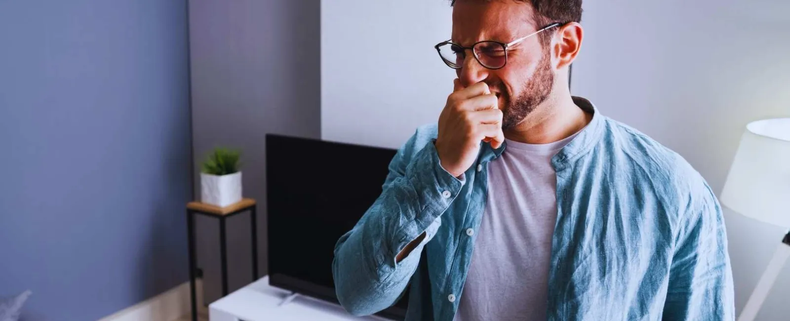 Man with glasses sneezing or coughing in modern living room with TV and lamp in background