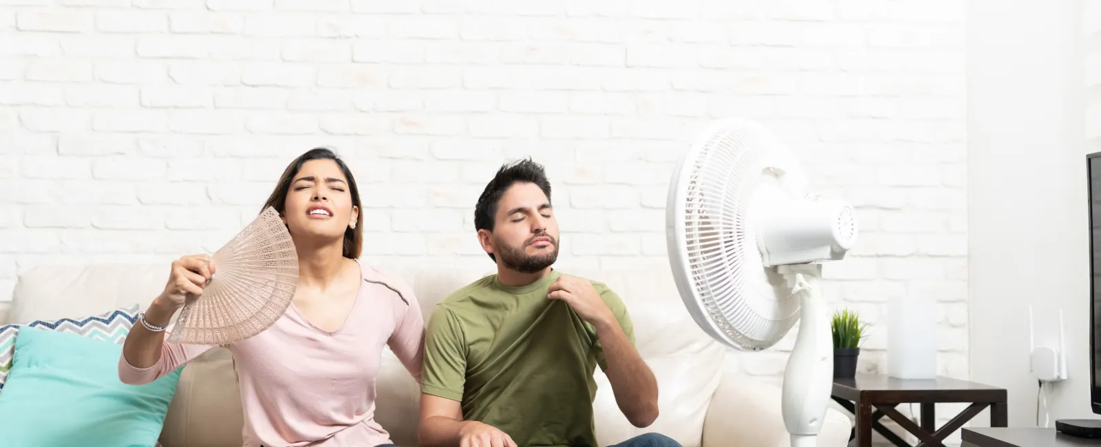Young man and woman sitting on sofa using fan and hand fan to cool off in hot room.