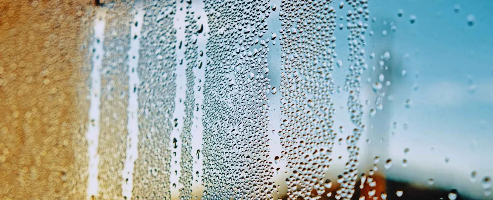 Close-up of water droplets on a window glass with blurred outdoor background in warm and cool tones.
