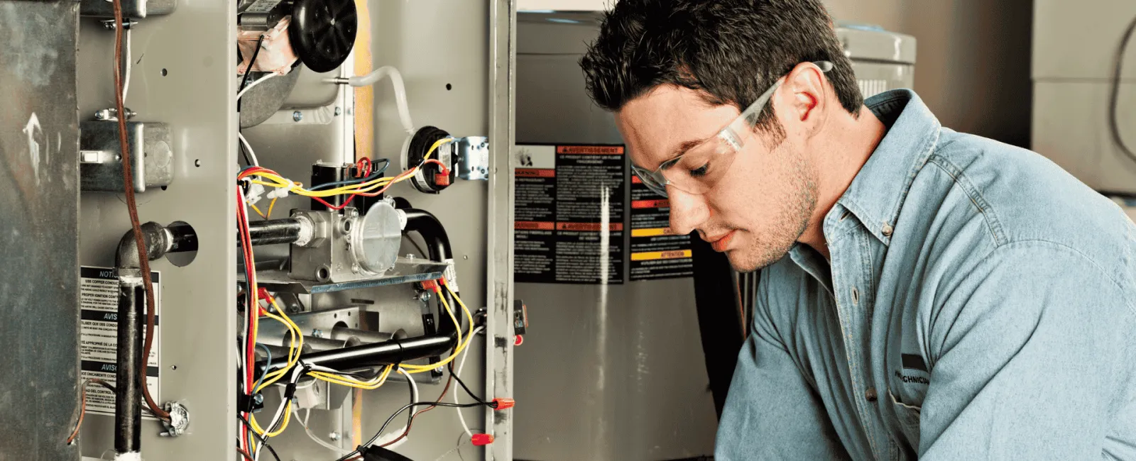 Technician wearing safety glasses inspecting furnace wiring with a multimeter in a mechanical room.