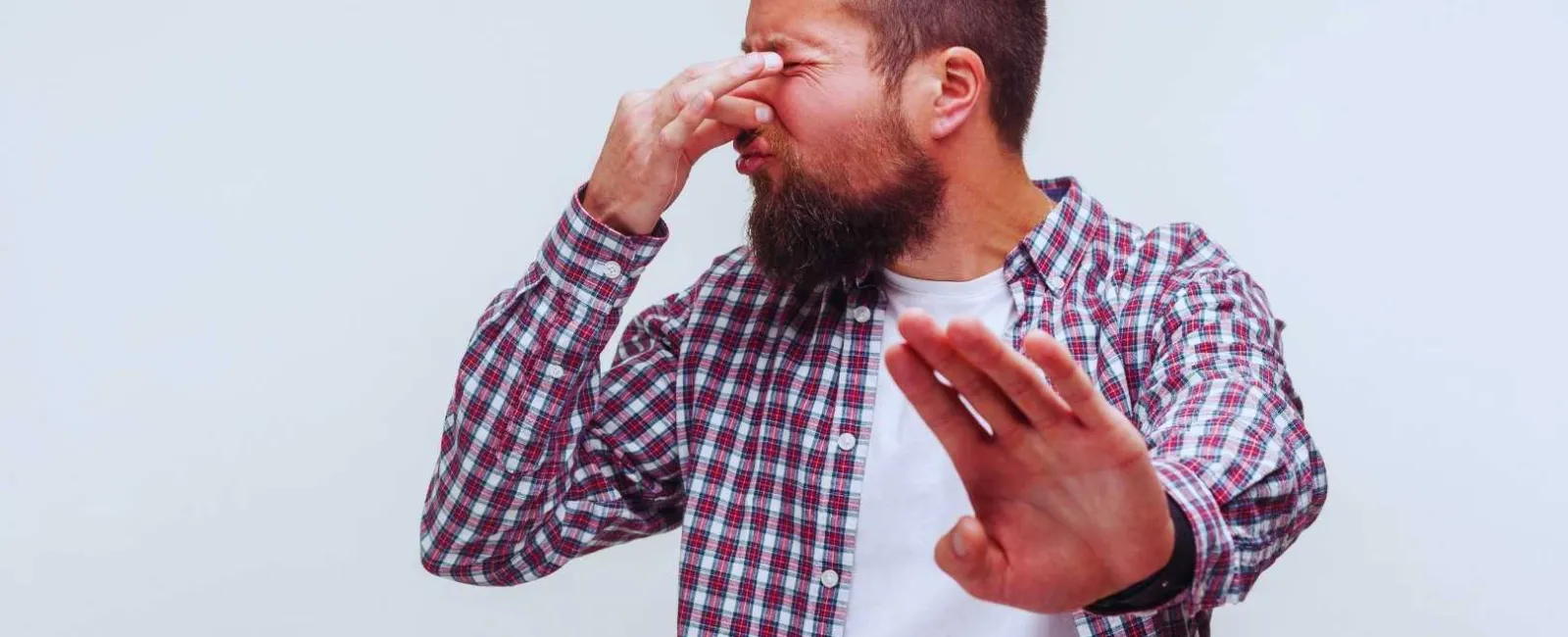 Man in plaid shirt pinching nose and raising hand, reacting to a bad smell or unpleasant odor.