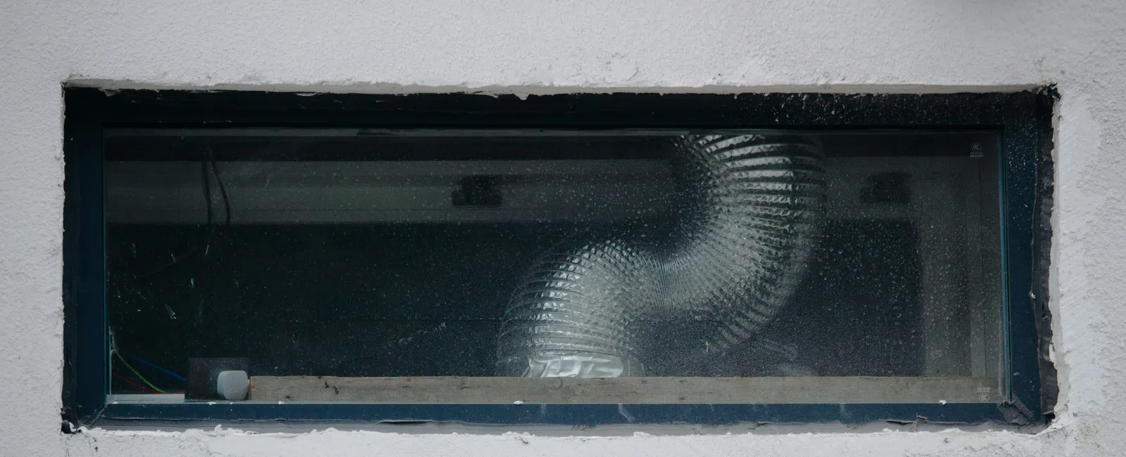 Small rectangular basement window with a ventilation duct visible through the glass in a white wall.