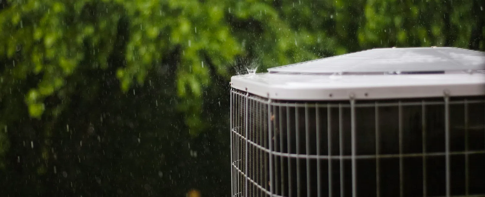 Outdoor air conditioning unit in rain with blurred green trees in background, showing water droplets falling.