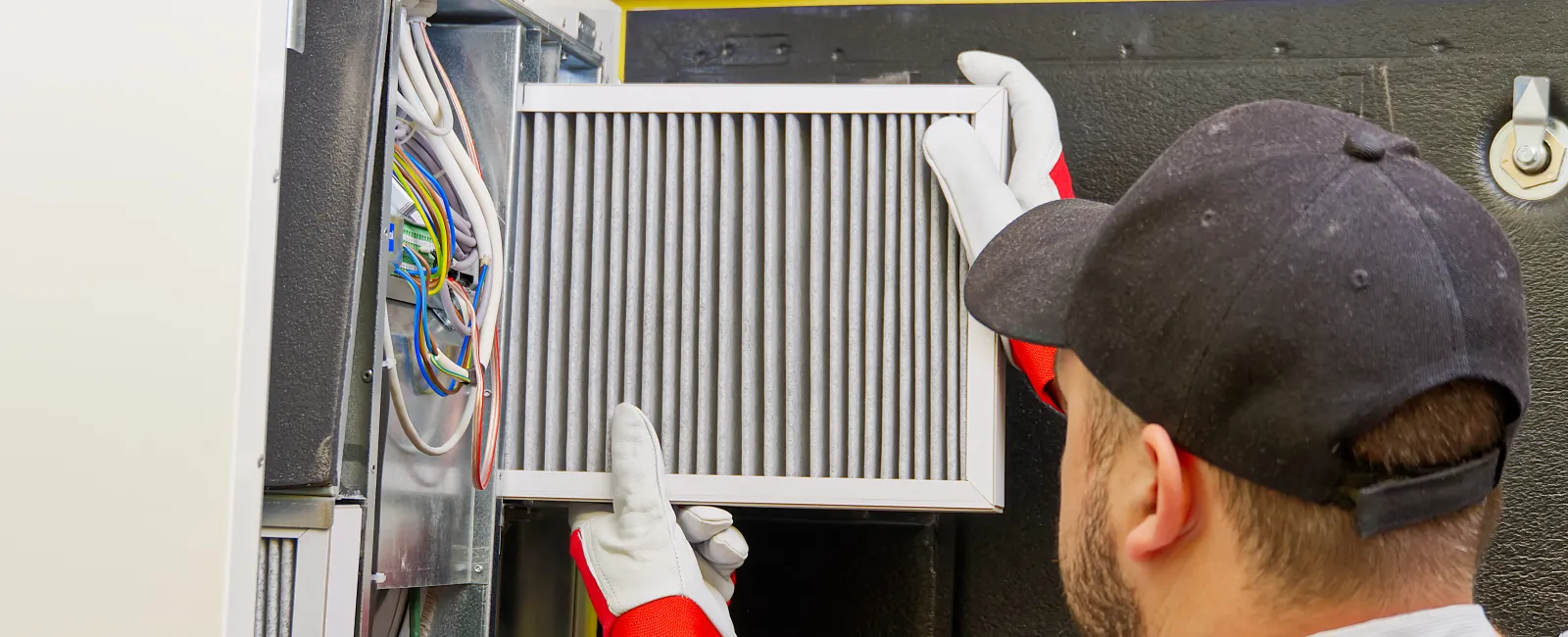 Technician replacing an air filter in a furnace with tools and gloves in a well-lit room.