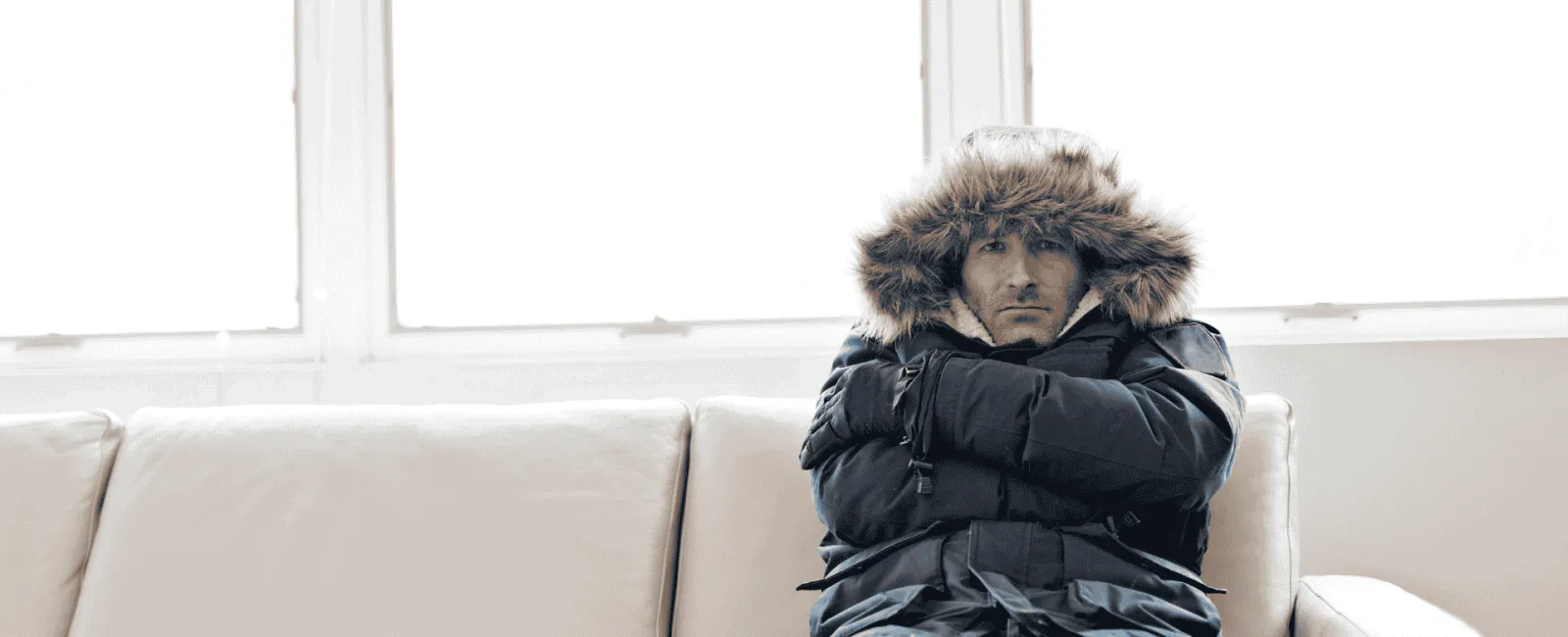 Man dressed in winter jacket sitting on a beige sofa indoors with arms crossed, looking cold and uncomfortable.