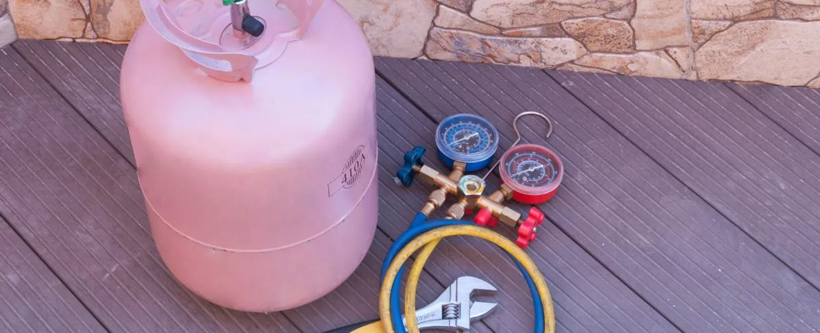 Pink refrigerant gas cylinder with pressure gauges, hoses, and adjustable wrench on wooden floor against stone wall.