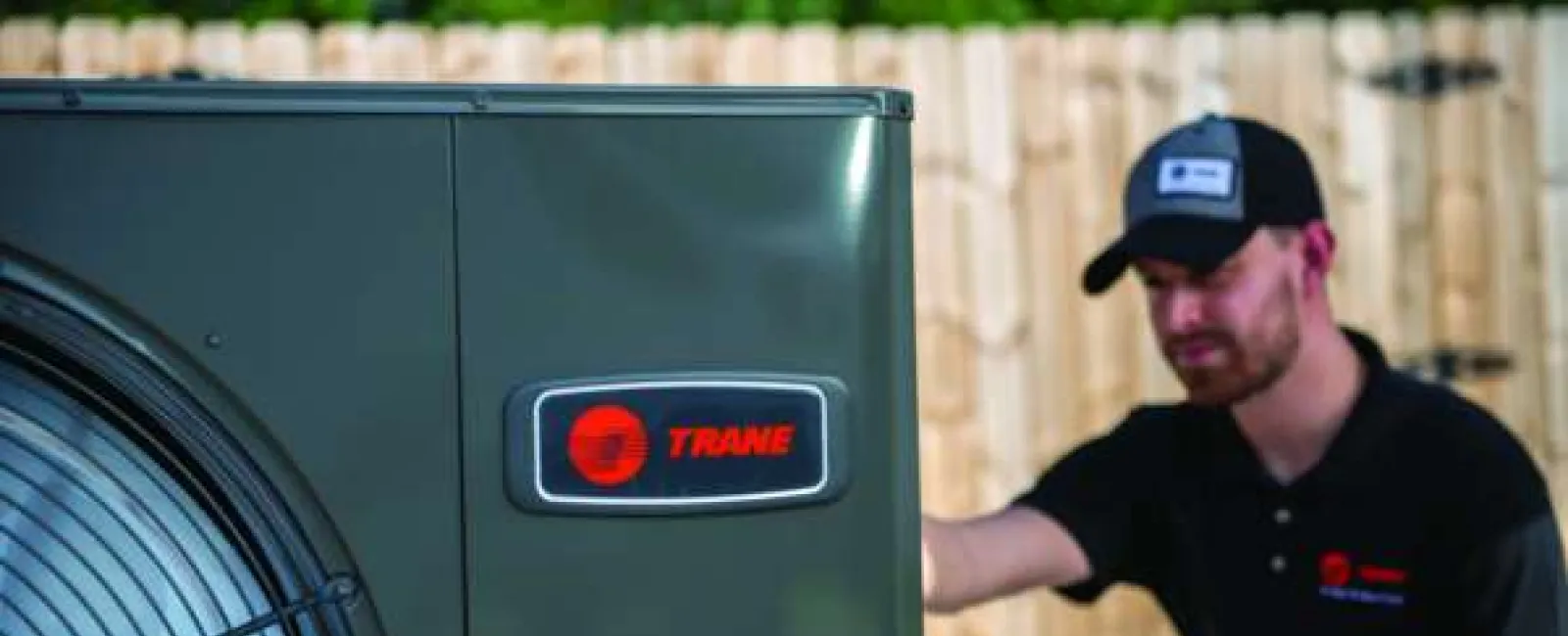 Technician inspecting a Trane HVAC unit outdoors in front of a wooden fence during daytime.