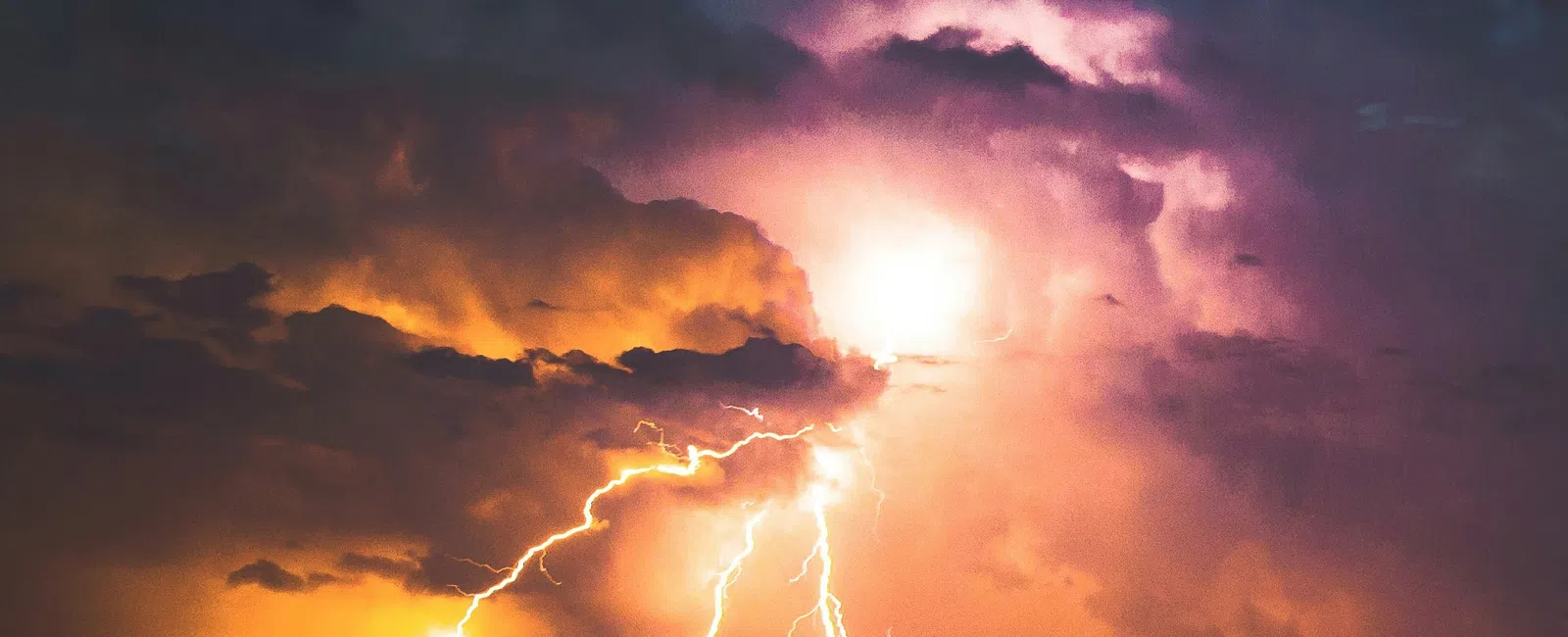 Multiple lightning bolts illuminating dark clouds over silhouetted trees at night with an orange and purple sky.