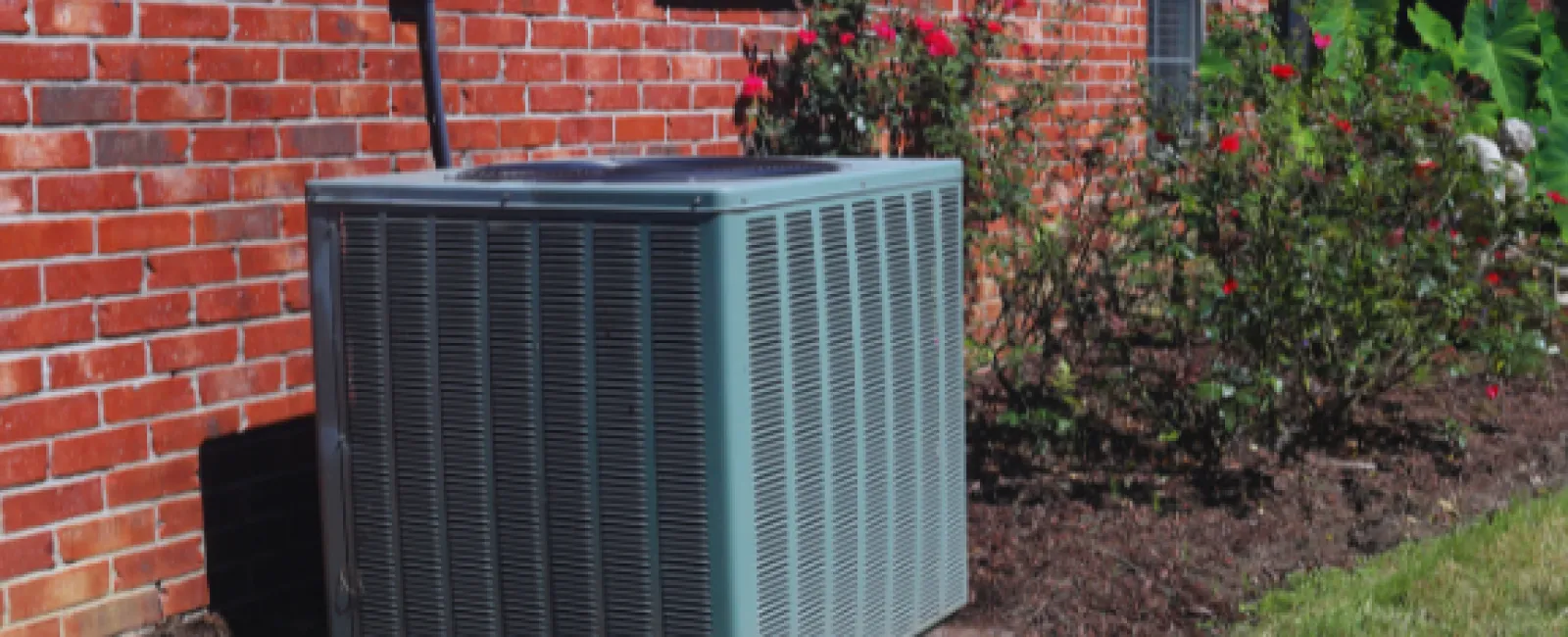 Outdoor air conditioning unit installed beside a brick house with a garden of flowering plants nearby.