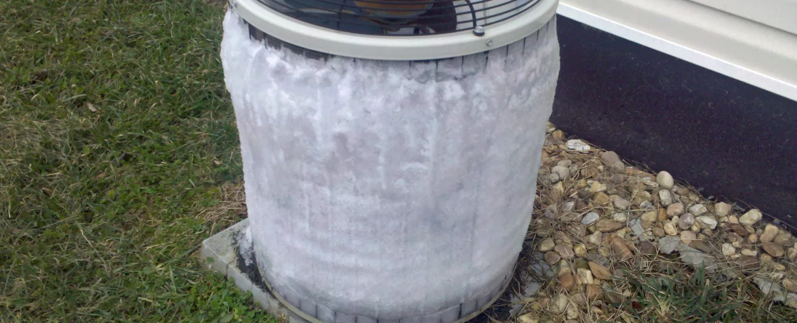 Outdoor air conditioning unit covered in ice and frost due to freezing temperatures next to house siding.