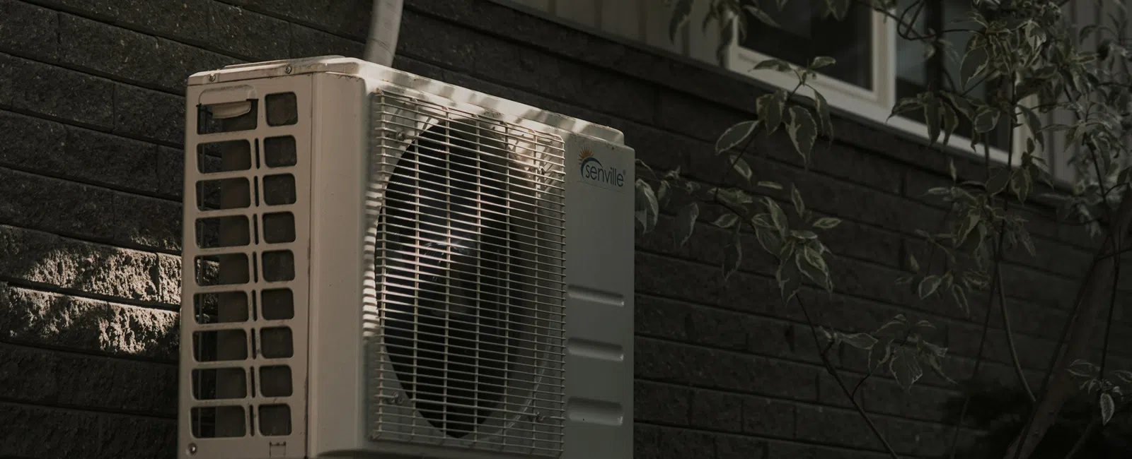 Outdoor air conditioning unit mounted on a stone wall outside a dark brick house with surrounding foliage.