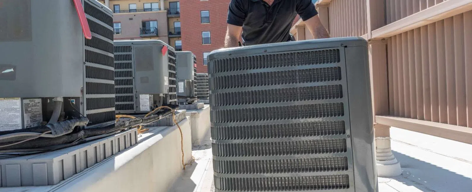 Technician in hard hat adjusting rooftop HVAC unit on sunny day near apartment building.