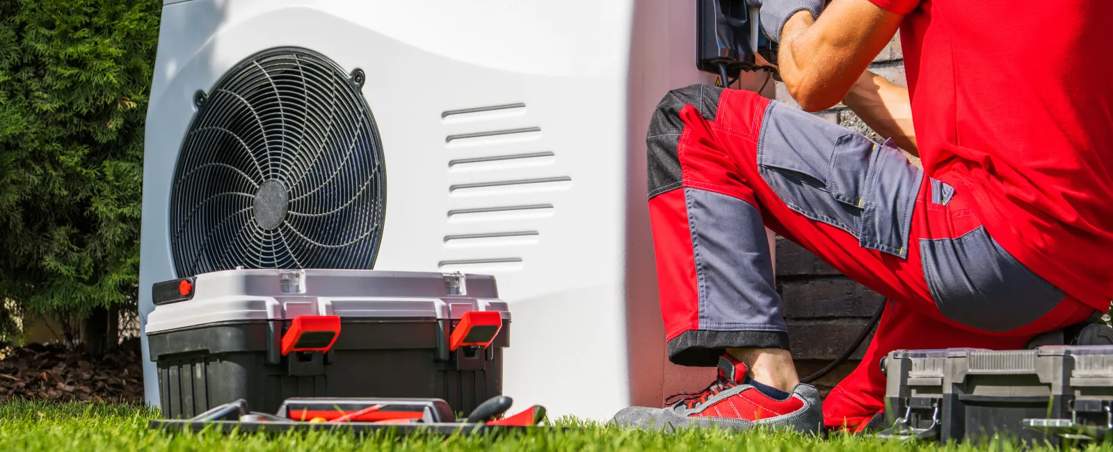 Technician in red uniform repairing or installing an outdoor air conditioning unit on a green lawn.