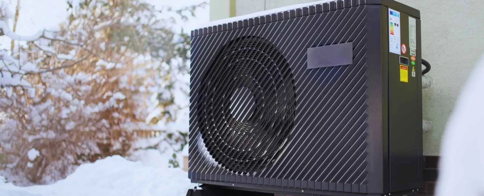 Outdoor heat pump unit installed on blocks beside a house in snowy winter conditions with frosted bushes in background.