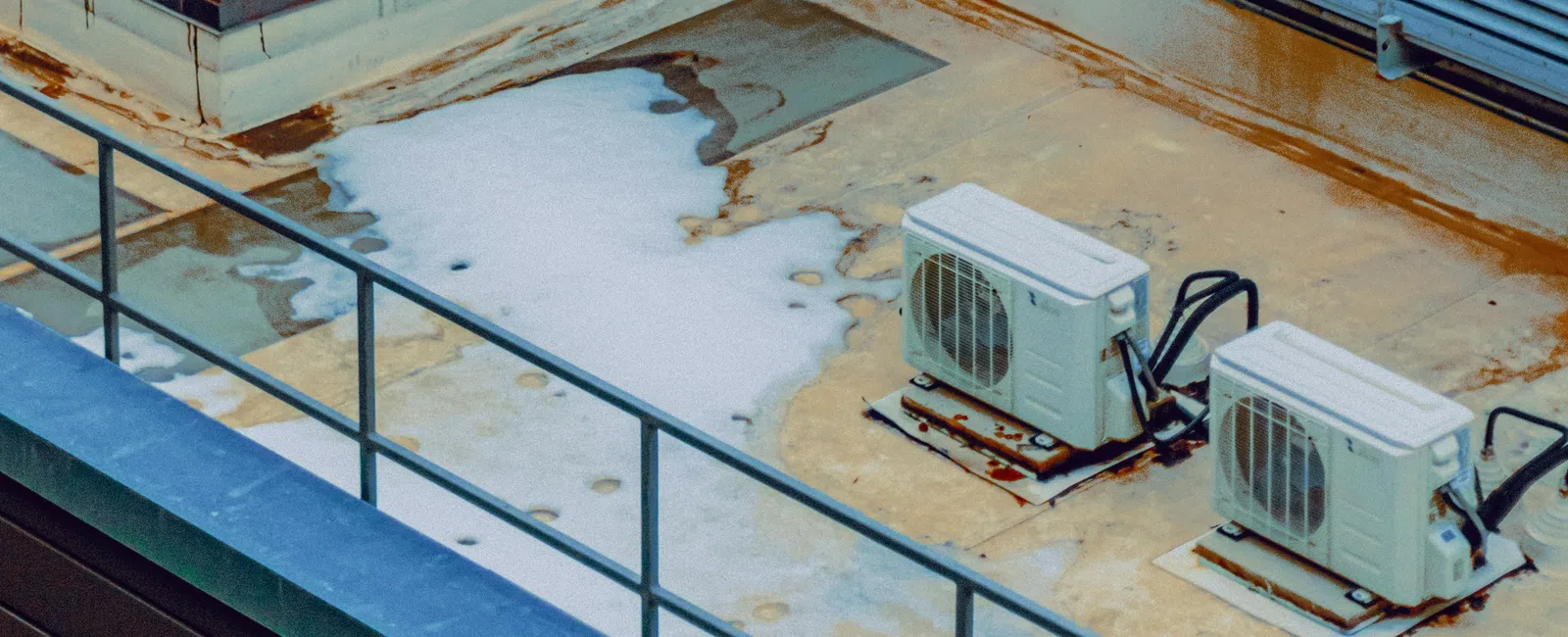 Industrial rooftop showing HVAC units, metal ladder, brick wall, and weathered concrete surfaces with some snow patches.