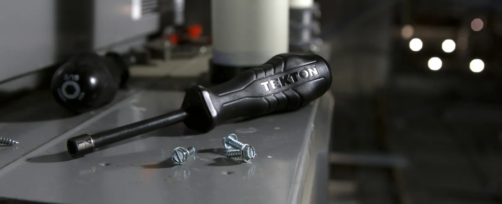 Close-up of a Tekton screwdriver and screws on a metallic surface in a workshop or industrial setting.