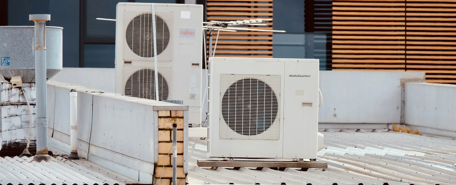 Air conditioning units installed on a rooftop against a building with wooden blinds on windows.