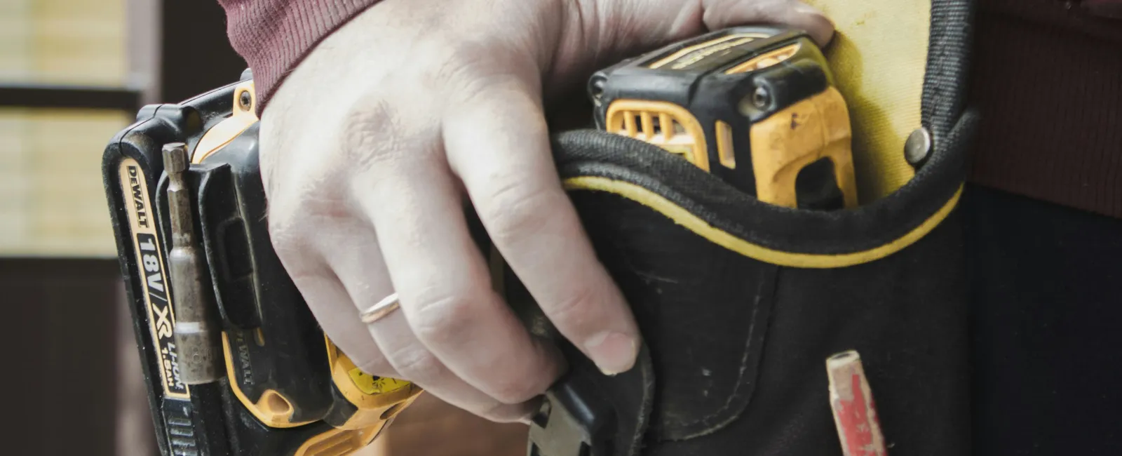 Close-up of a worker's hand holding a power drill with a tool belt against a wooden wall background.