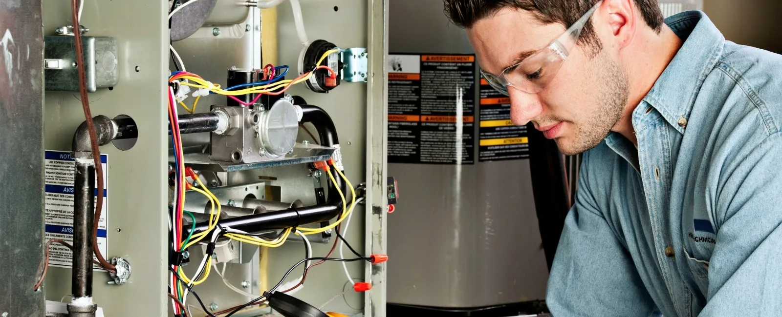 Technician wearing safety glasses inspecting and repairing electrical wiring inside a home furnace unit.