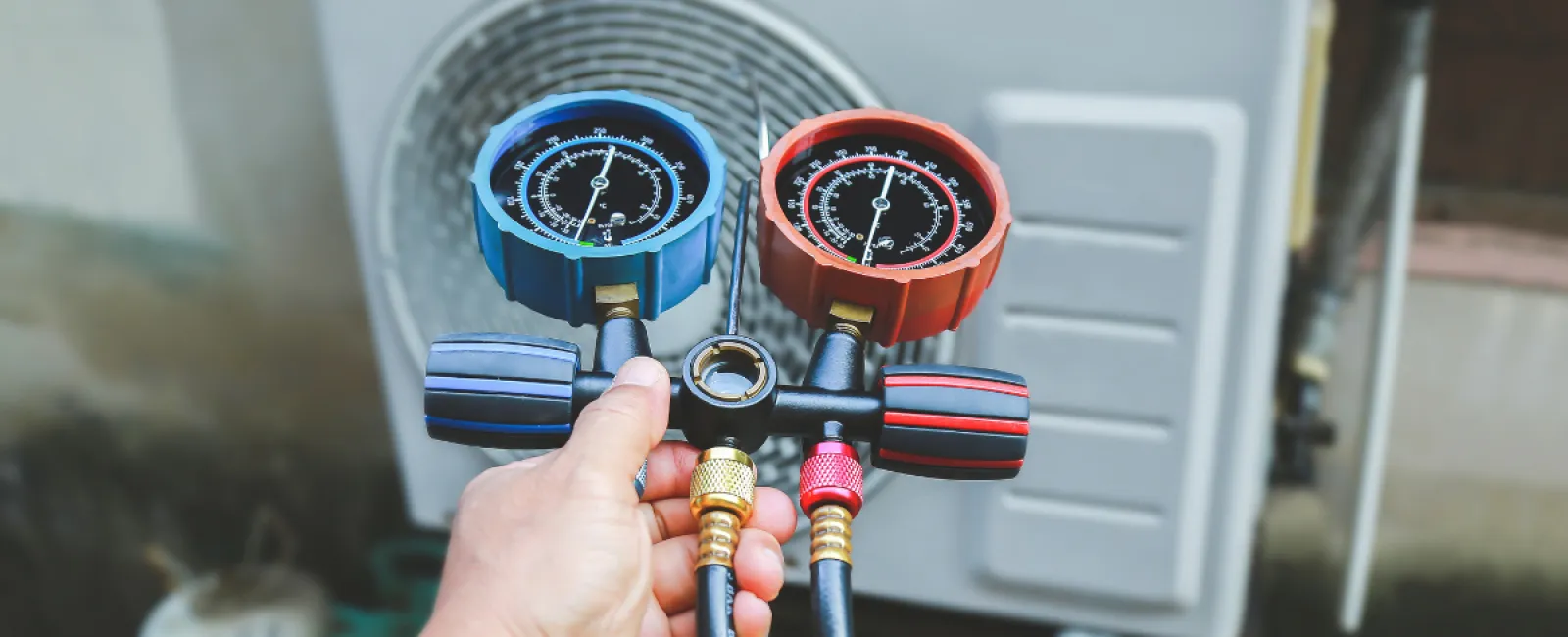 Hand holding HVAC manifold gauges with blue and red dials in front of an air conditioning unit outdoors.