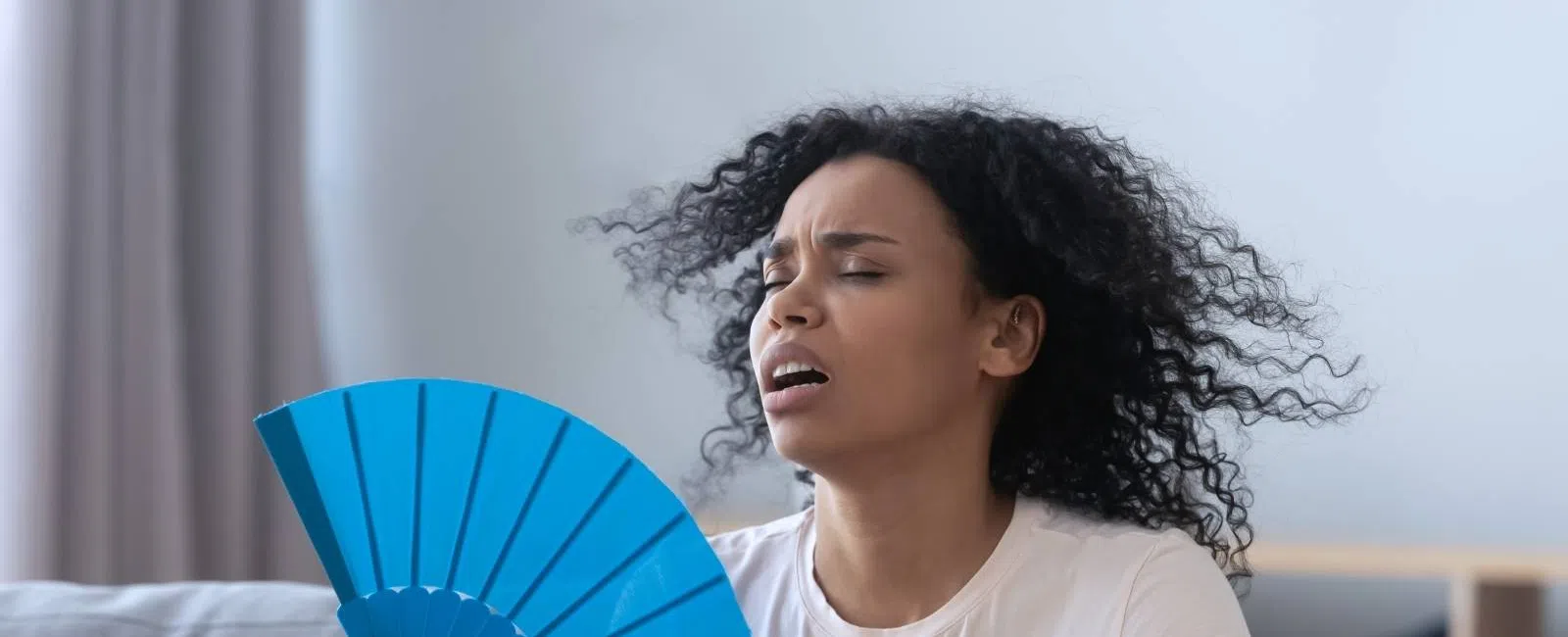 Woman in a white shirt fanning herself with a blue hand fan, appearing hot and uncomfortable indoors.