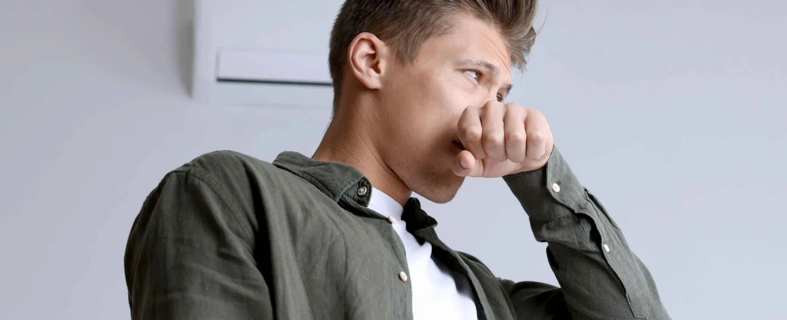 Young man covering cough with hand indoors wearing casual clothes under air conditioner unit.