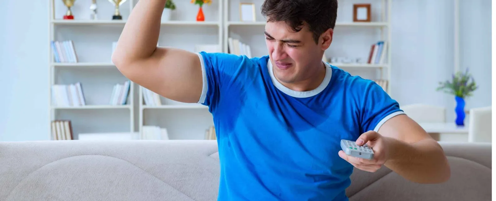Young man in blue shirt flexing his bicep and holding a TV remote on a couch at home.