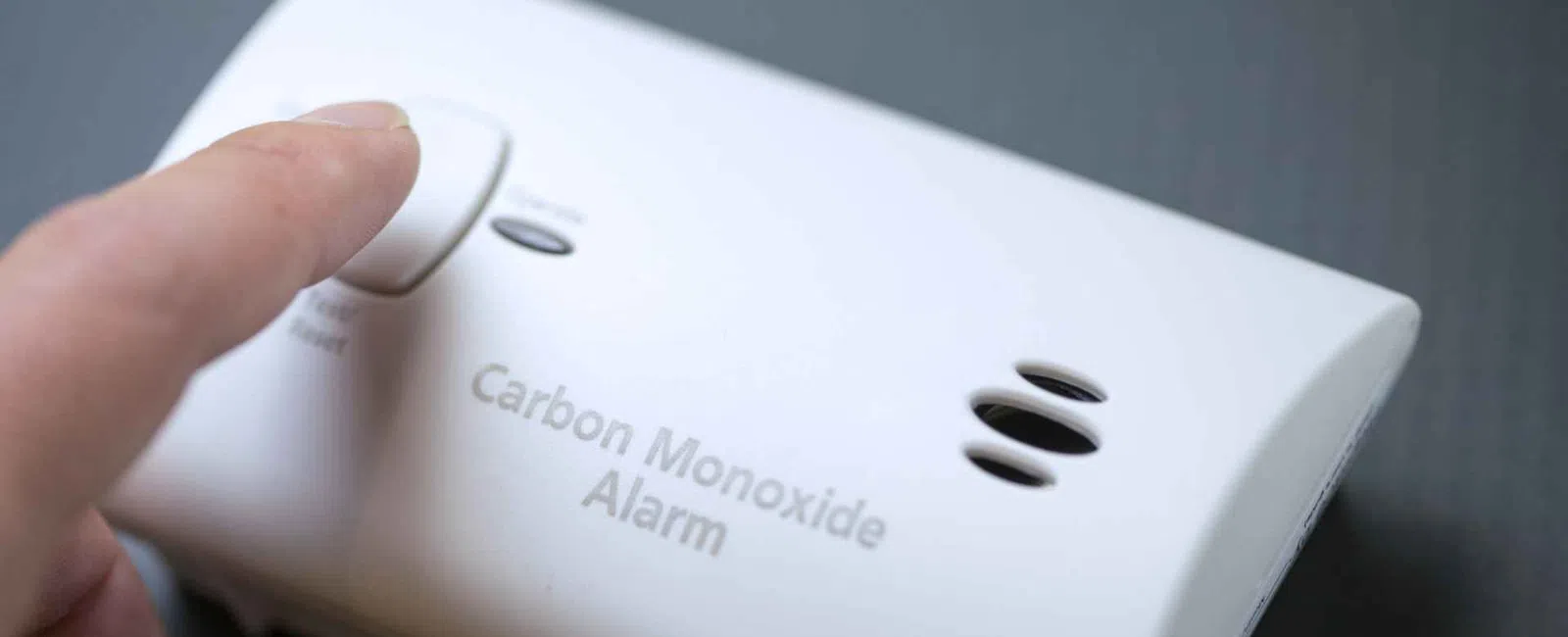 Finger pressing test button on white carbon monoxide alarm mounted on a wall.