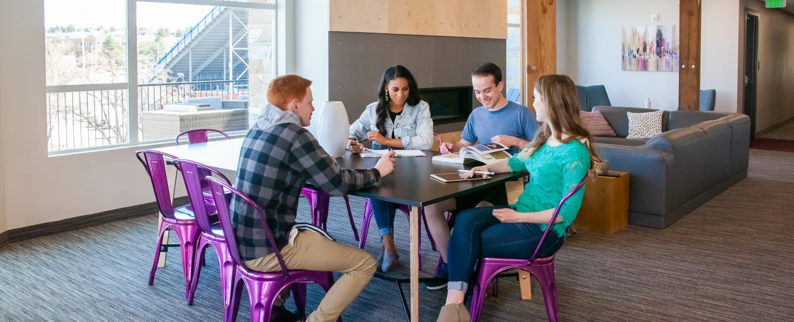 Four young adults sitting on purple chairs around a table studying together in a bright, modern lounge area.