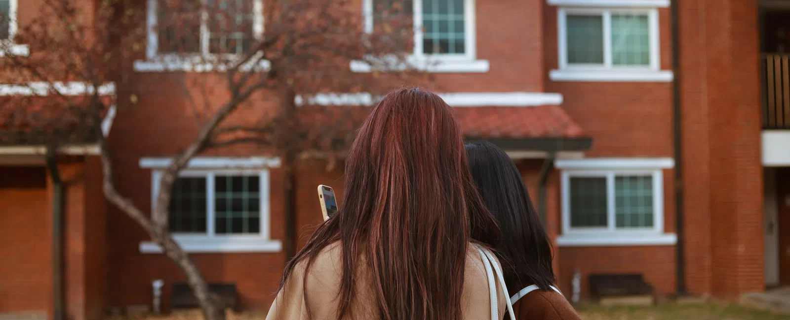 Two women with long hair stand outside a brick apartment building, one holding a smartphone and a white bag.