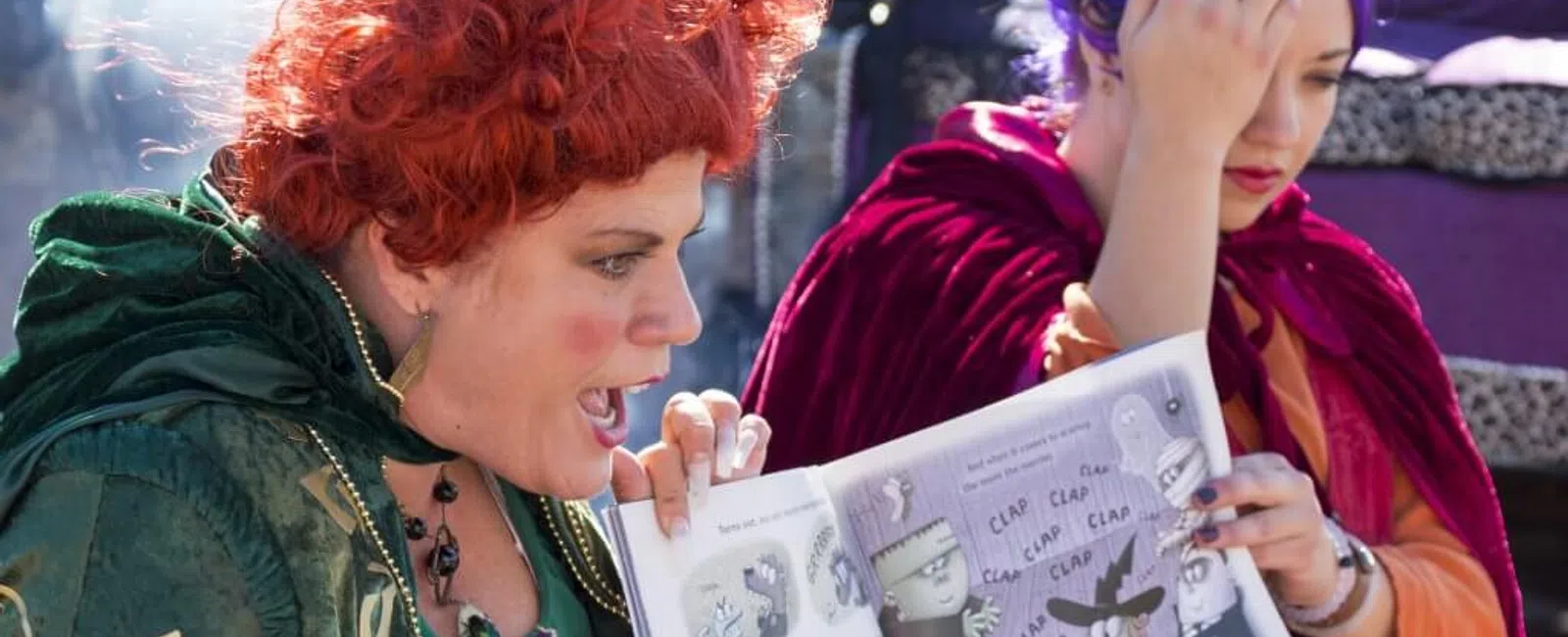 Two women in costumes animatedly reading a storybook outdoors, surrounded by a playful atmosphere.