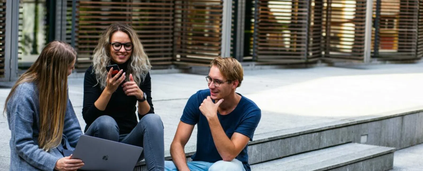 Three friends engaged in conversation on outdoor steps, sharing moments with a laptop and smartphone.