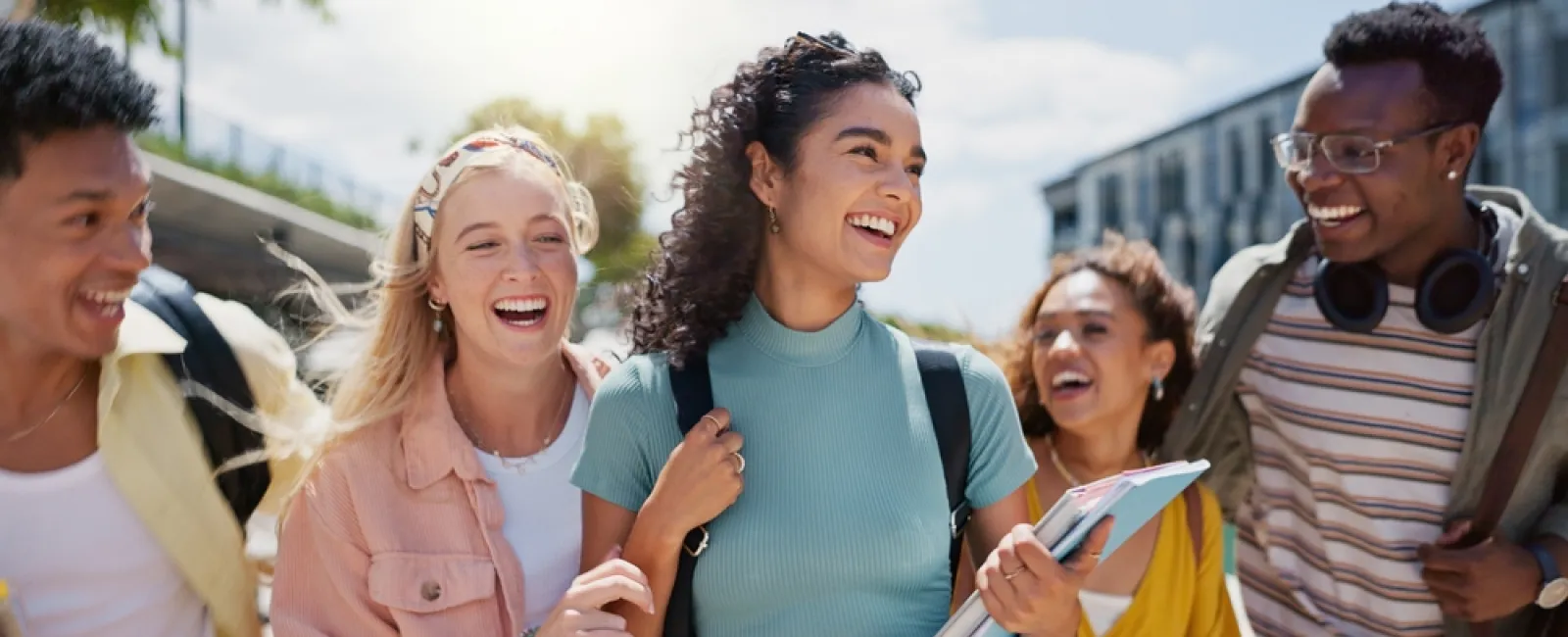 Group of diverse young adults smiling and walking outdoors on a sunny day with books and backpacks.